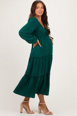 woman wearing a dark green maternity dress with a tiered skirt and long sleeves, posing with her baby bump visible
