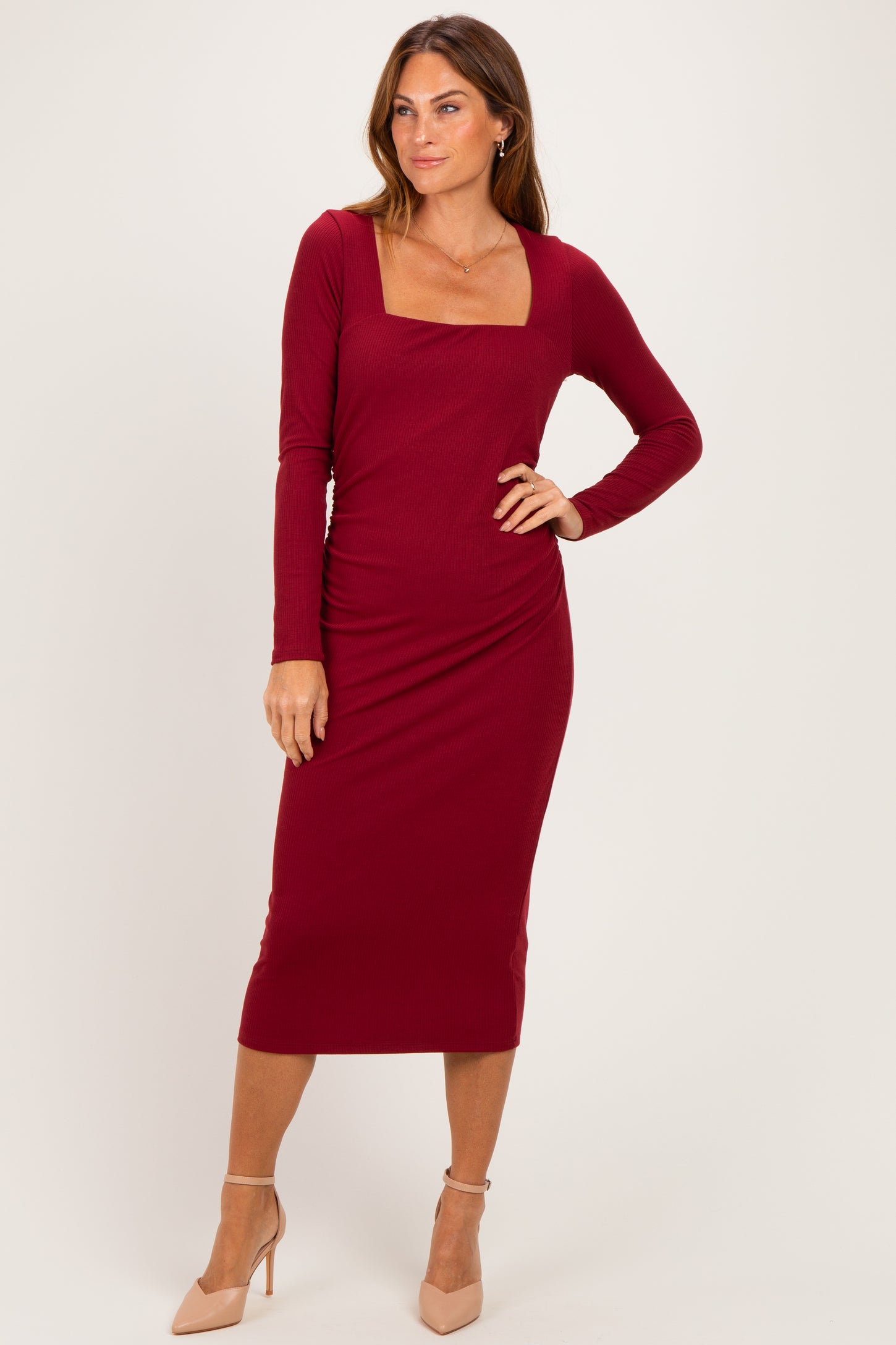 woman wearing a burgundy long sleeve midi dress with a square neckline and side ruching, standing in a neutral pose