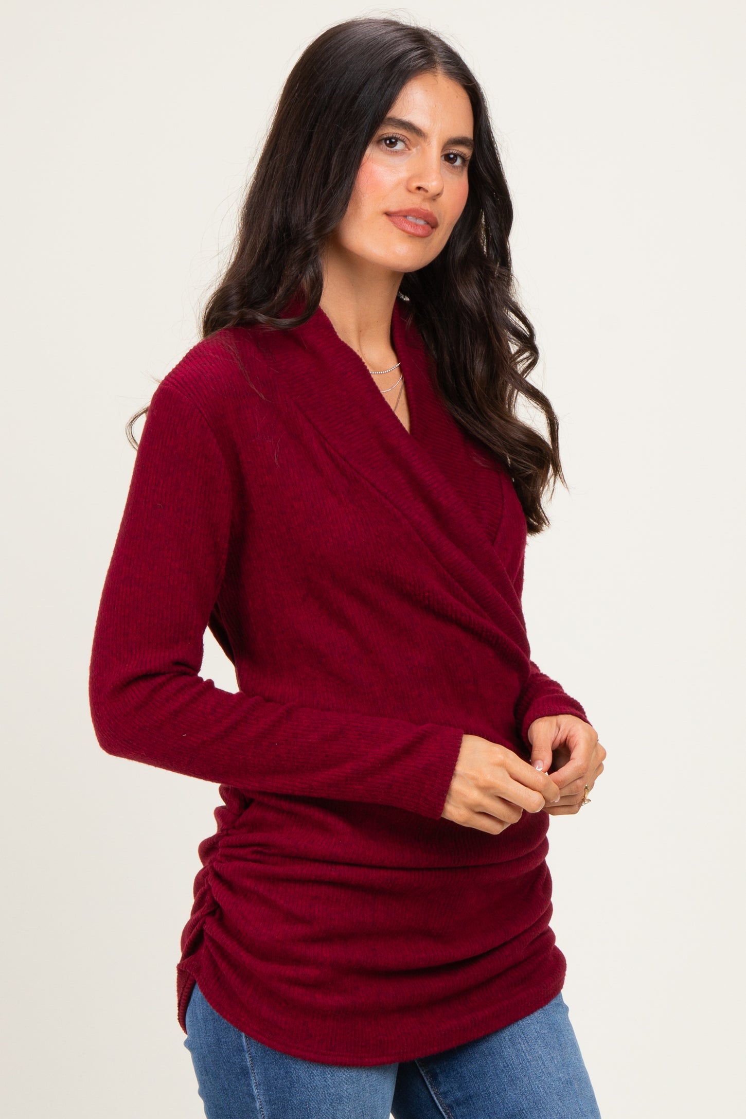 woman wearing a dark red wrap-style sweater with side ruching and a relaxed fit, paired with blue jeans