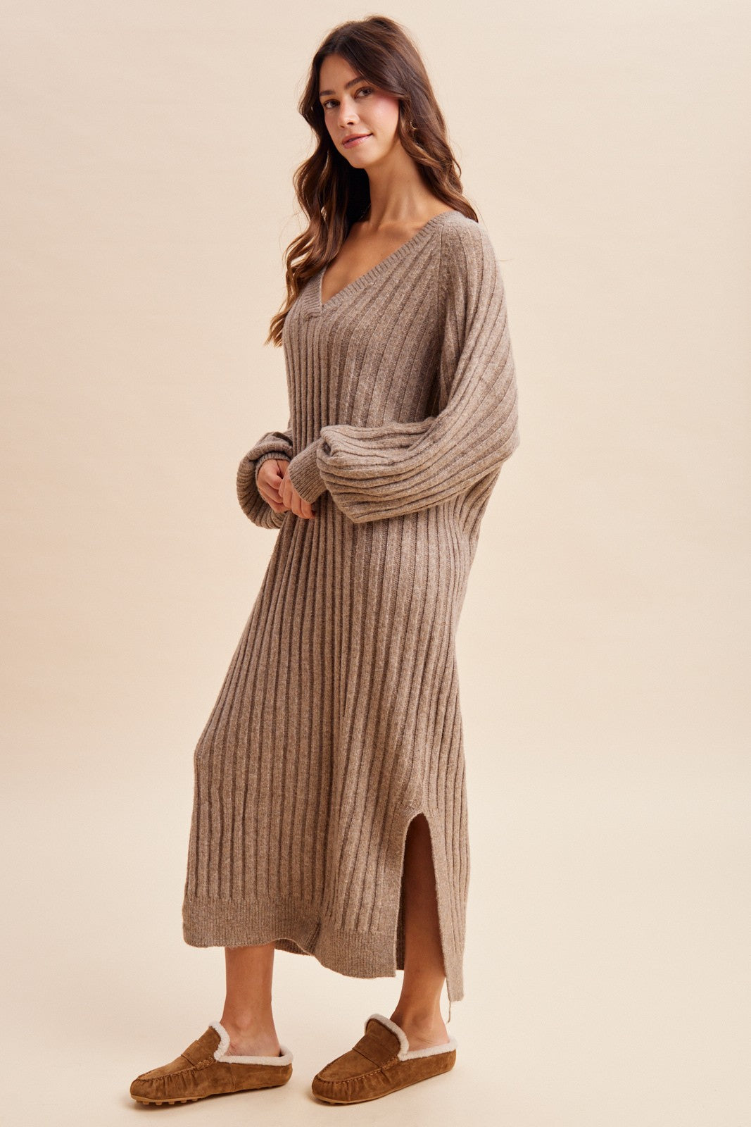 woman wearing a beige long sleeve ribbed sweater dress with side slits and styled with brown slippers