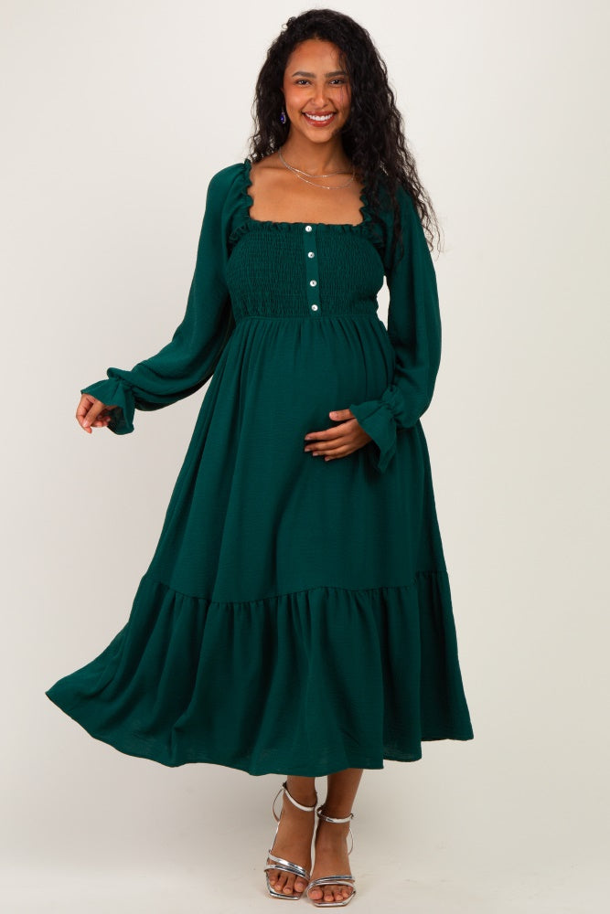 woman wearing a dark green maternity dress with a smocked bodice, long sleeves, and a tiered skirt, holding her baby bump