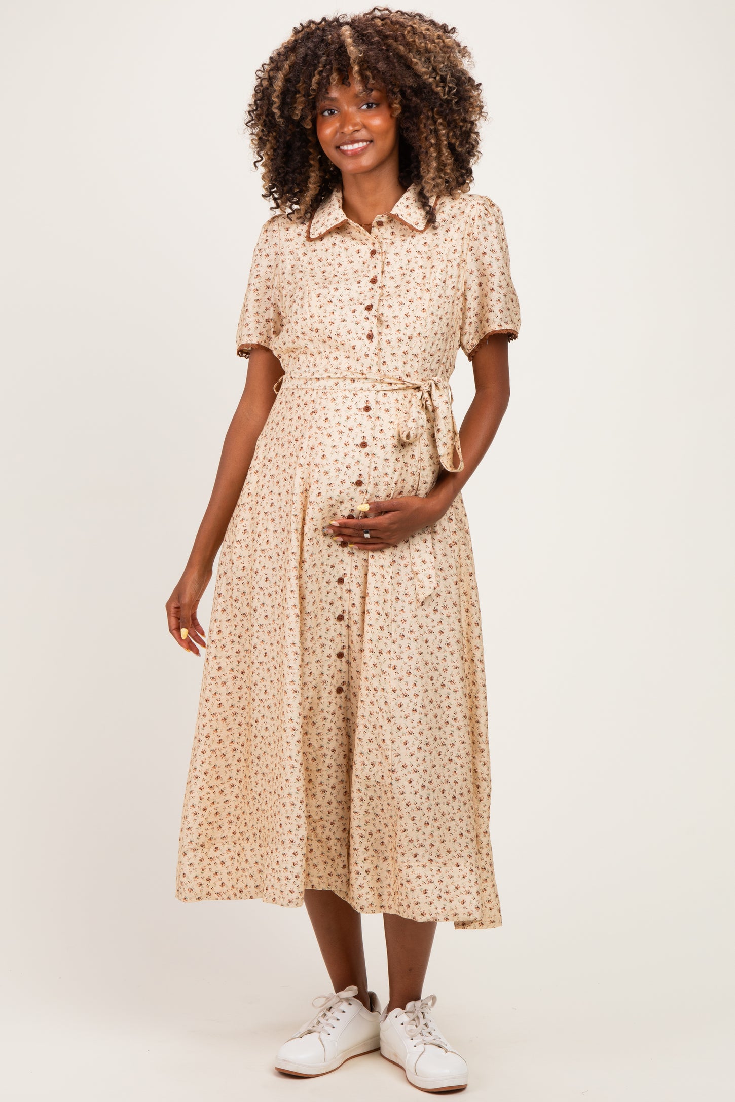 woman wearing a beige floral midi maternity dress with buttons and a tie waist, holding her baby bump