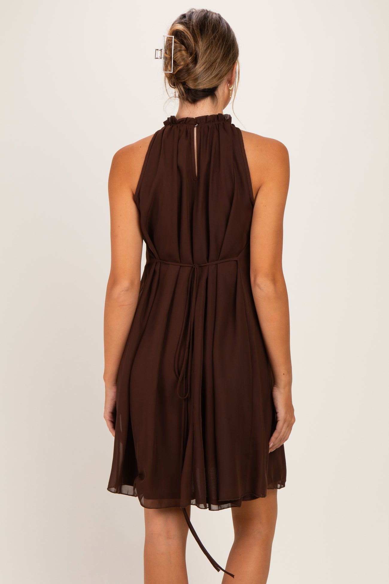 woman wearing a brown sleeveless maternity dress with a ruffled neckline and adjustable tie waist from the back