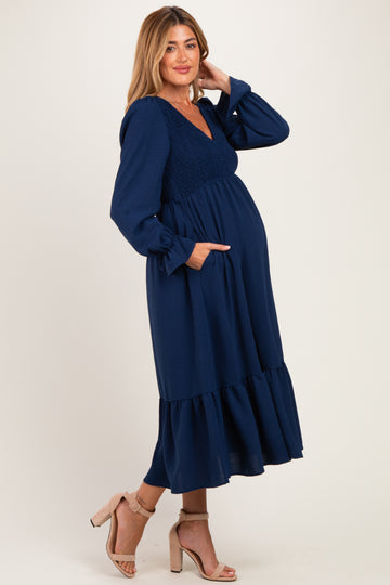 woman wearing a navy blue maternity dress with long sleeves, smocked bodice, and tiered skirt, showcasing her baby bump
