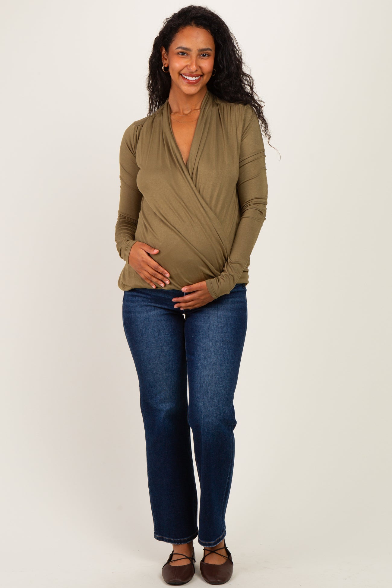 woman wearing an olive green wrap maternity top and blue jeans, cradling her baby bump