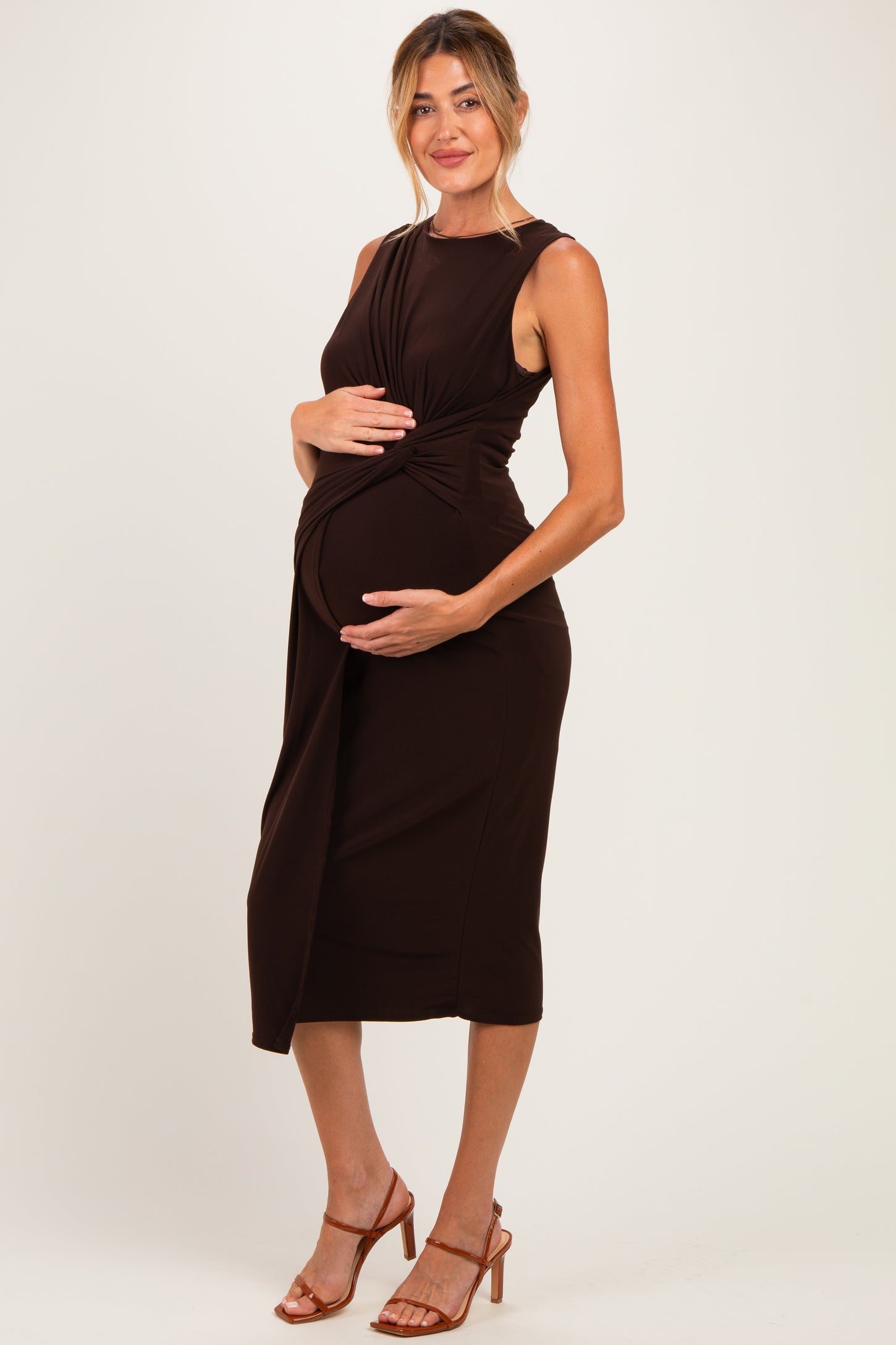 woman wearing a brown sleeveless maternity dress with a twisted waist and holding her baby bump