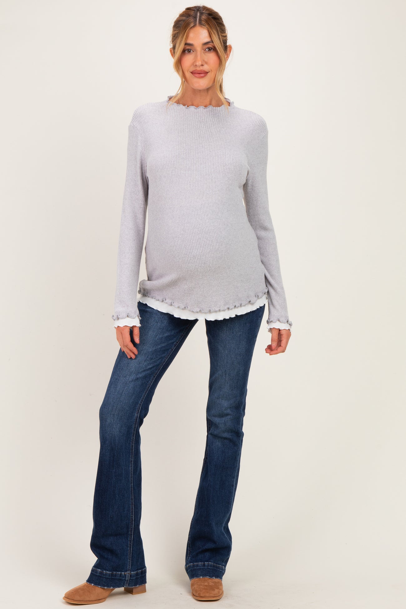 woman wearing a light gray maternity sweater with scalloped hem and long sleeves, paired with blue flared jeans and a visible baby bump