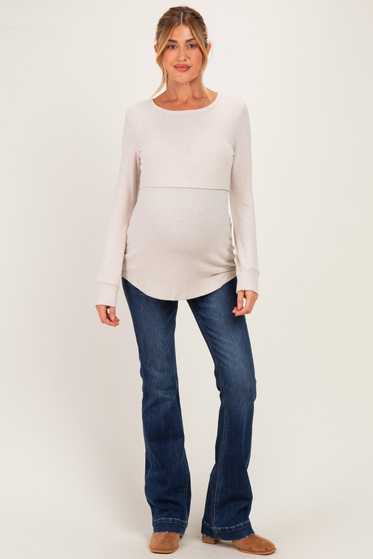 woman wearing a light beige long sleeve maternity top and blue flared jeans, showing off her baby bump