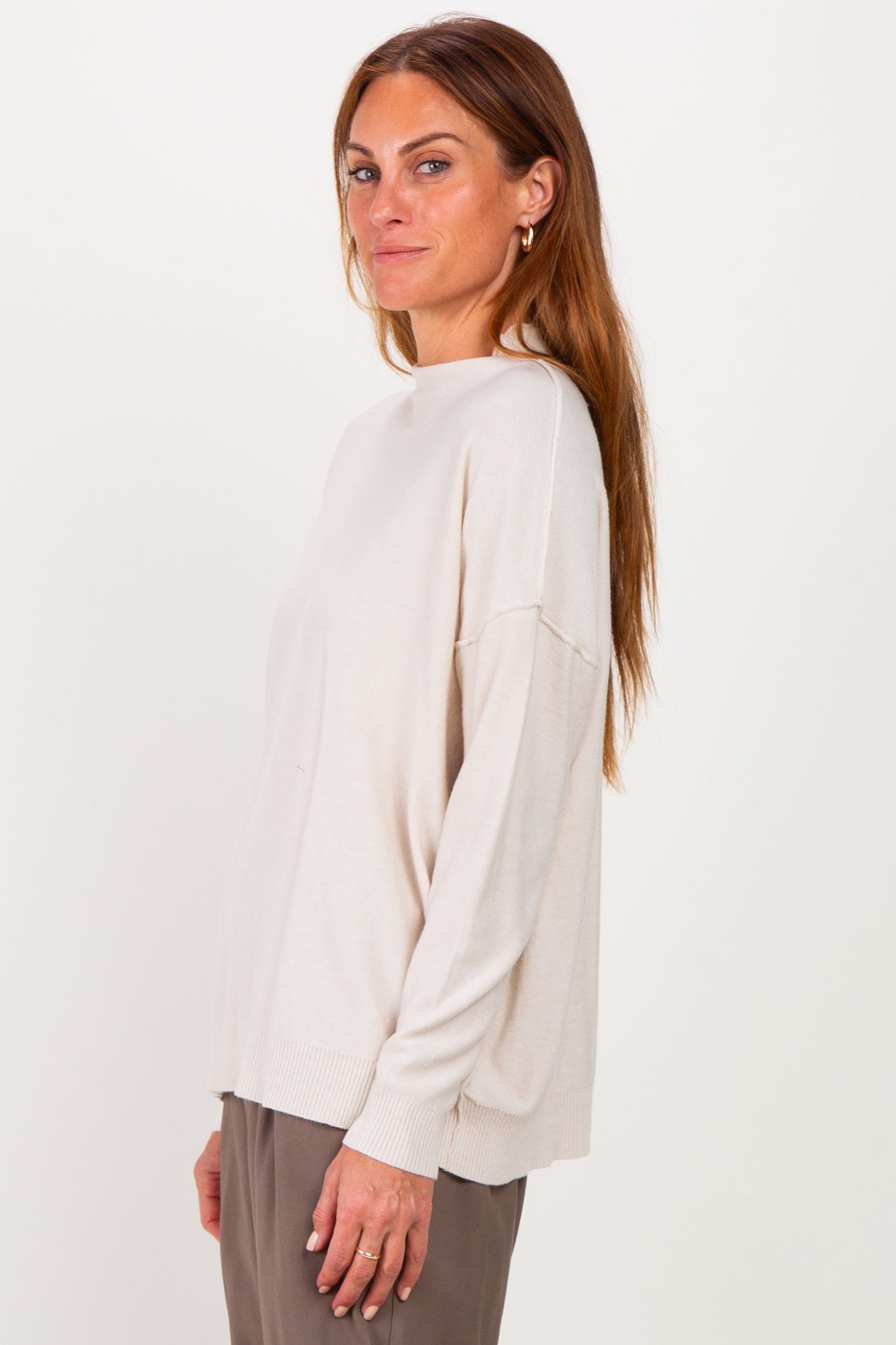 woman wearing a light beige oversized sweater with a high neckline and side slits, paired with brown trousers