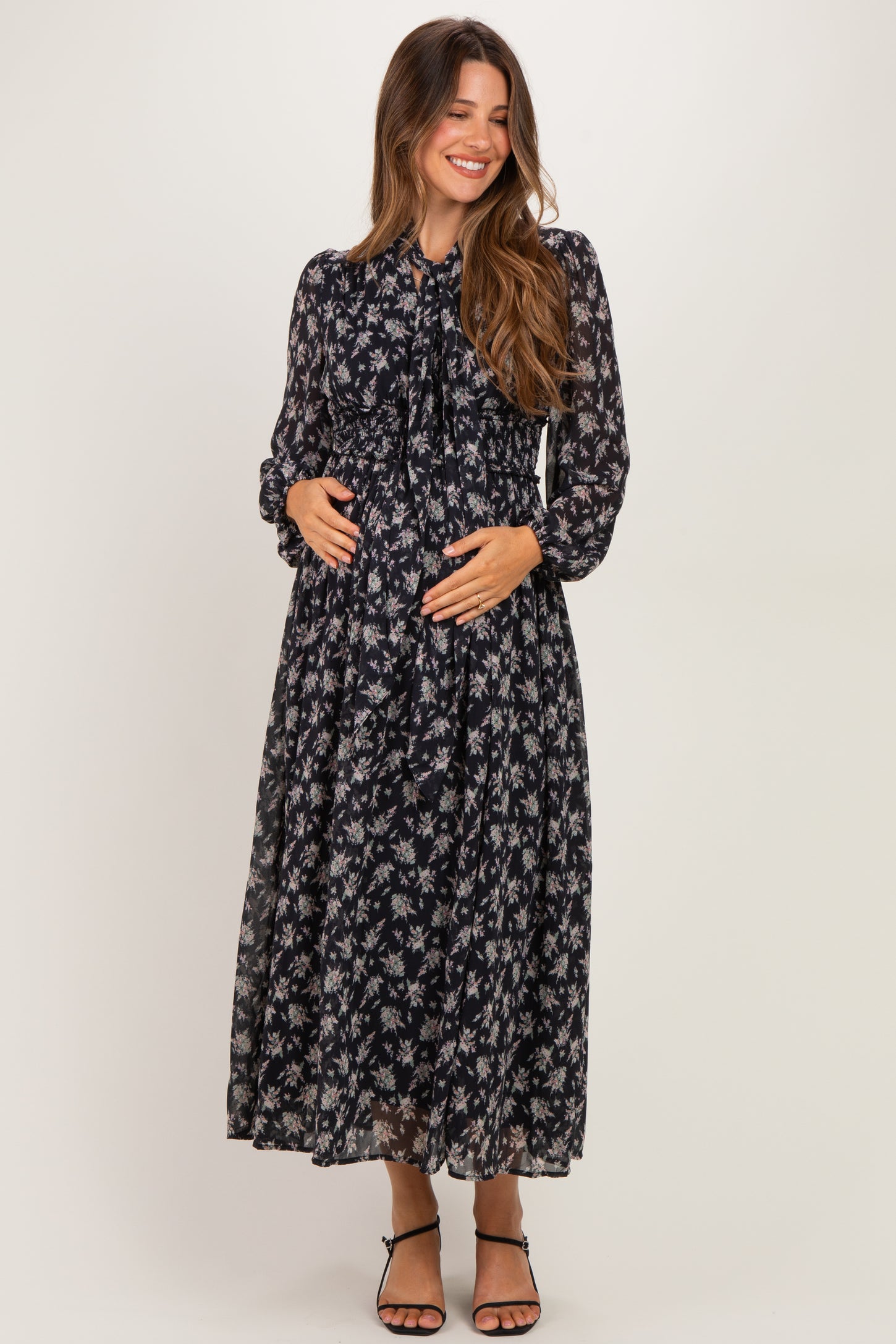 woman wearing a navy floral maternity maxi dress with long sleeves and a tied neckline, holding her baby bump