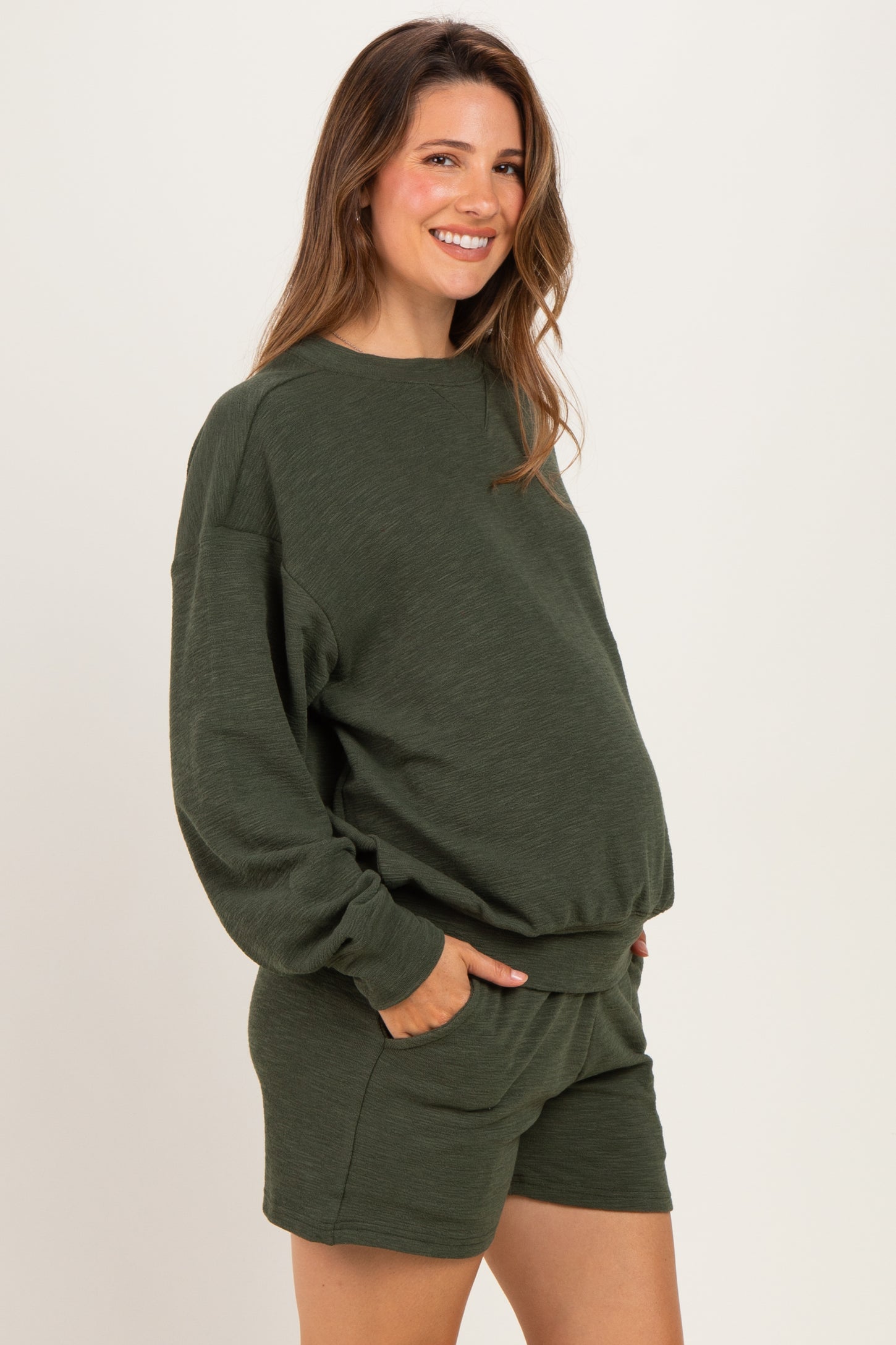 woman wearing a dark green maternity sweatshirt and matching shorts, showing her baby bump with pockets in both garments
