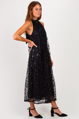 woman wearing a black sleeveless dress with intricate lace detailing and a flowing silhouette, paired with black heels