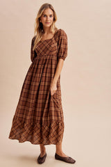woman wearing a long brown plaid maxi dress with puff sleeves and a tiered hem, standing against a neutral background