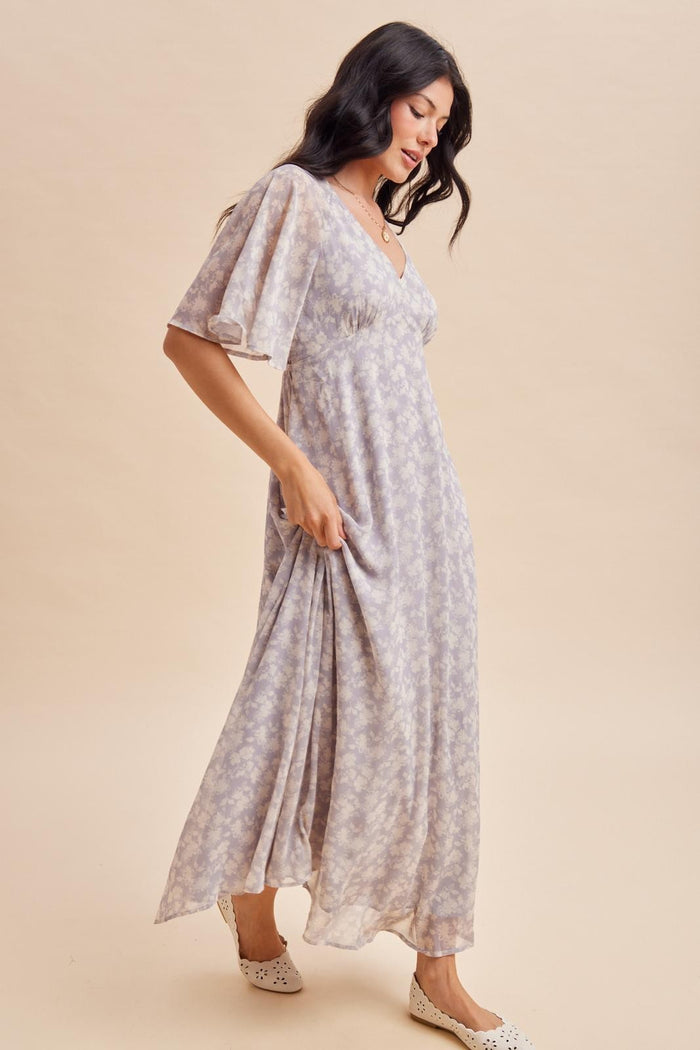 woman wearing a lilac floral maxi dress with flutter sleeves and a v-neckline, standing on a light background