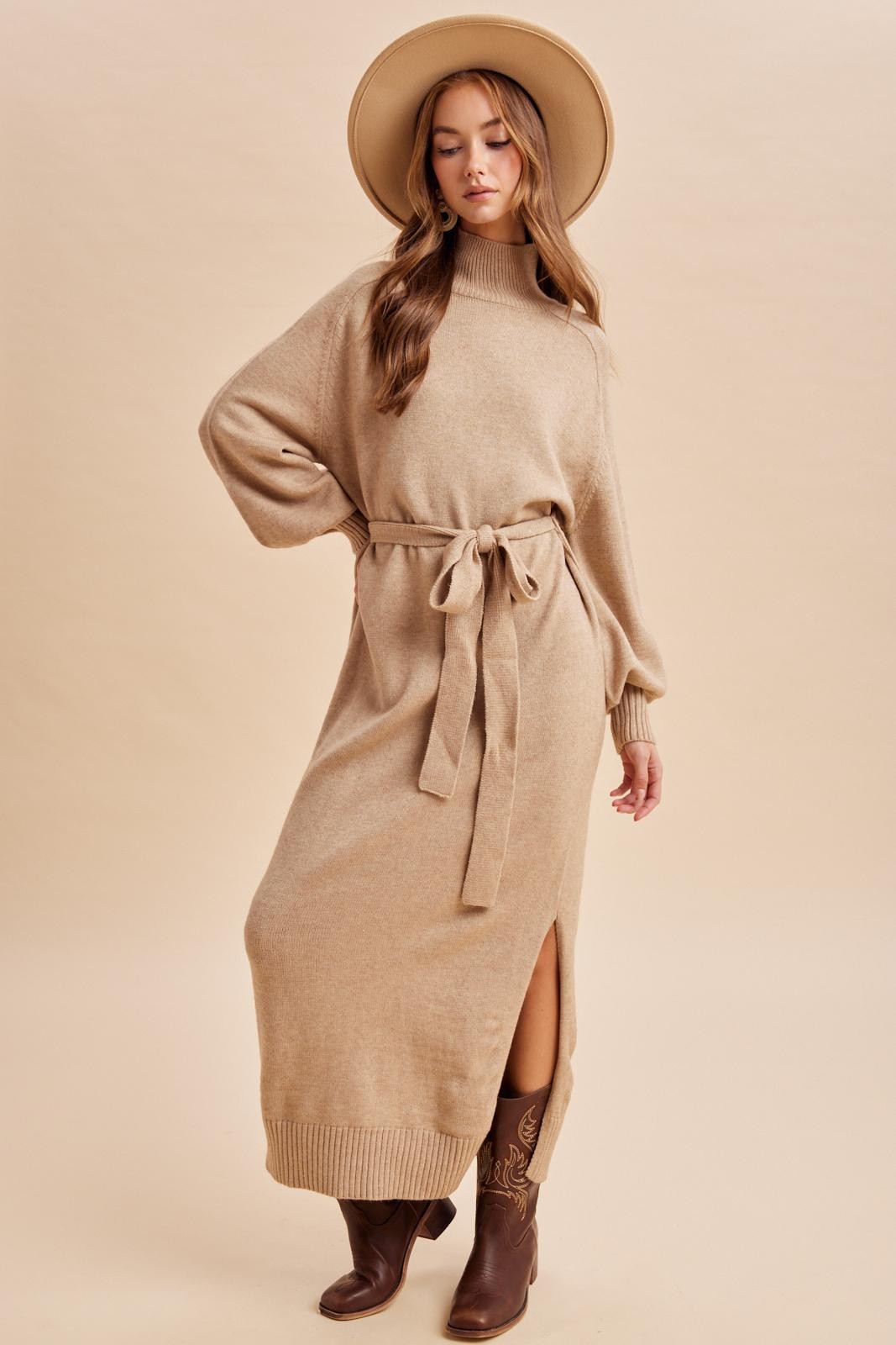 woman wearing a beige long sleeve belted sweater dress with a side slit and a wide-brimmed hat, paired with brown boots
