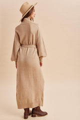 woman wearing a beige long sleeve cardigan dress with a belted waist and brown ankle boots, facing away from the camera
