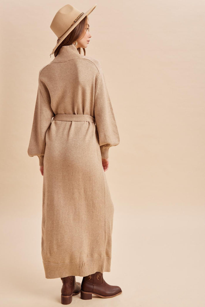 woman wearing a beige long sleeve cardigan dress with a belted waist and brown ankle boots, facing away from the camera