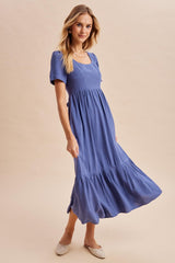 woman wearing a blue ribbed maxi dress with short sleeves and a tiered skirt, smiling and standing on a neutral background