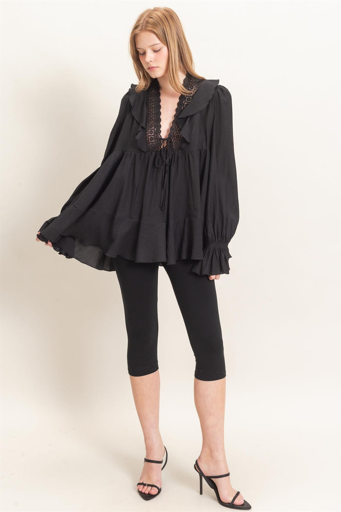 woman wearing a black oversized blouse with a ruffled collar and lace-up detail, paired with black leggings and heels
