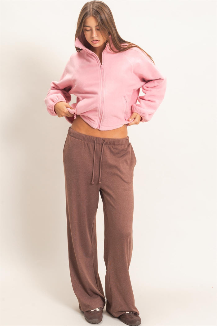 woman wearing a pink cropped fleece jacket and brown wide-leg pants, looking down while adjusting her outfit