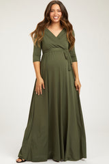 woman wearing a dark green maternity wrap maxi dress with three-quarter sleeves and a tied waist, standing and smiling
