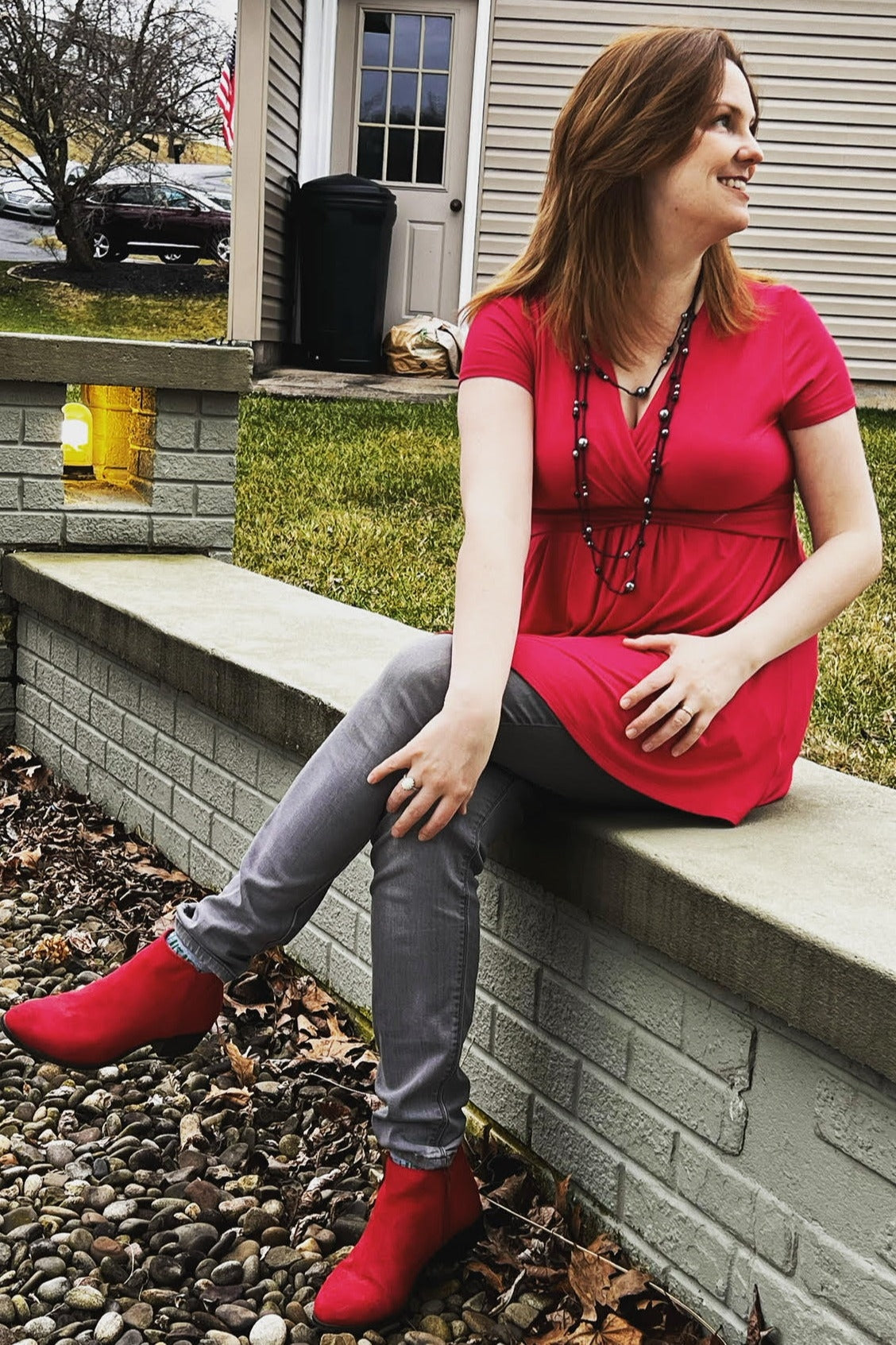 woman wearing a bright pink short sleeve tunic top, gray skinny jeans, and red ankle boots, sitting on a stone wall