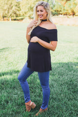 smiling pregnant woman in black off-the-shoulder maternity top and blue jeans standing in a grassy field