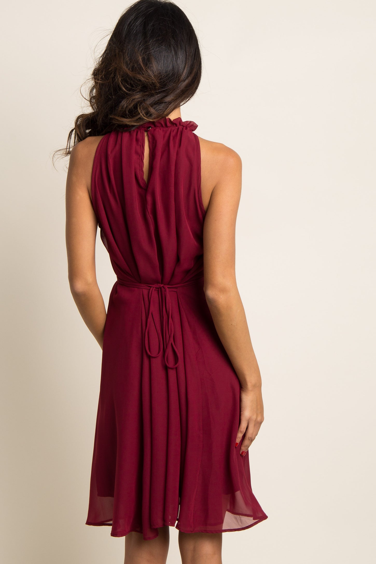 Back view of a woman wearing a sleeveless burgundy chiffon dress with a ruffled high neck and tie waist.