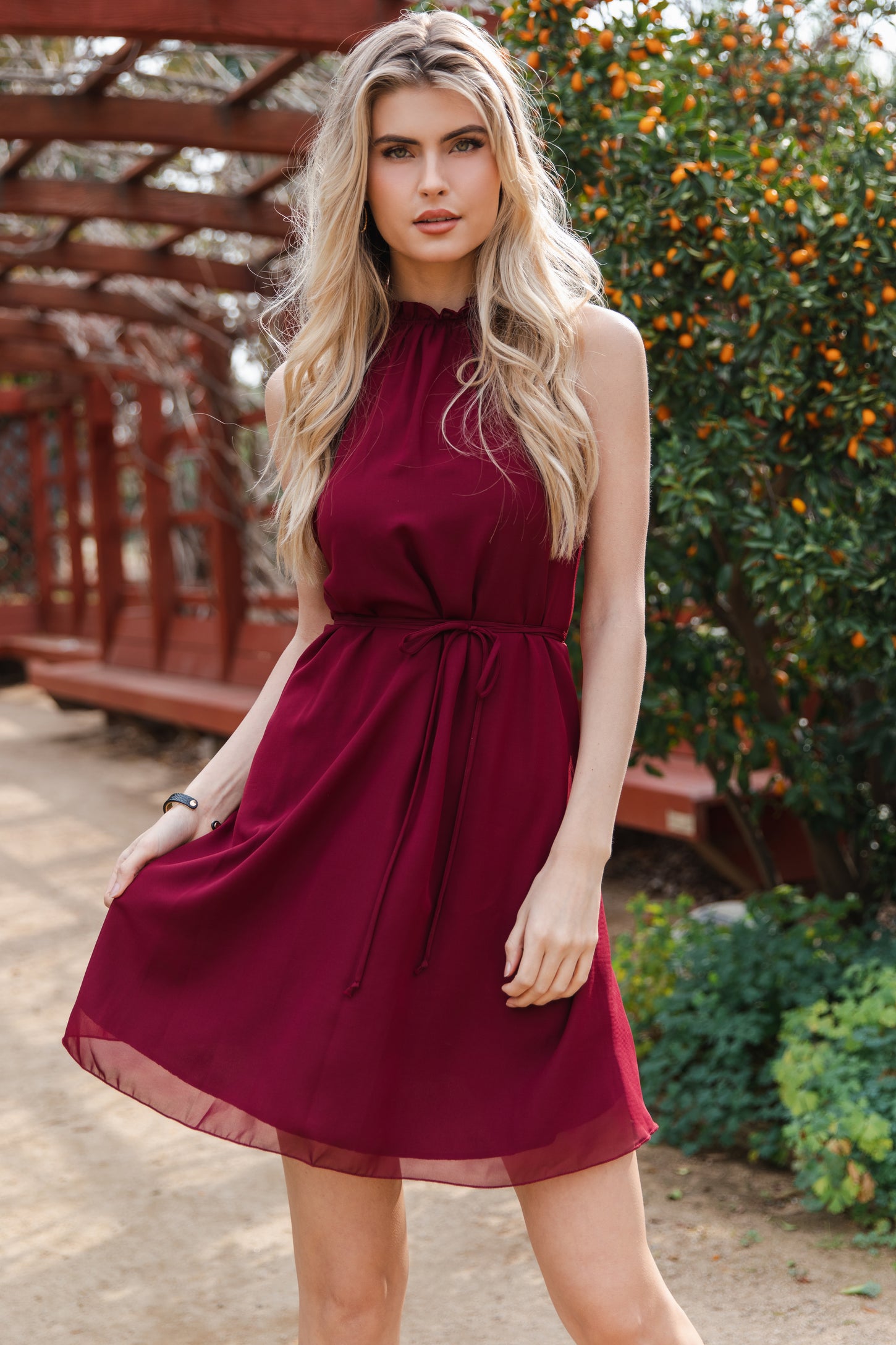 blonde woman wearing a sleeveless maroon halter dress with a tie waist in an outdoor garden setting