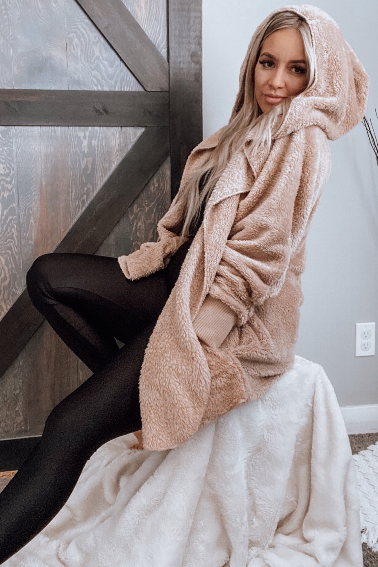 woman sitting on a stool wearing a beige oversized hooded teddy coat and black leggings