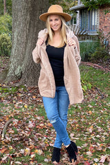 woman wearing a beige cozy faux fur jacket, black top, and jeans, smiling outdoors with a hat on her head