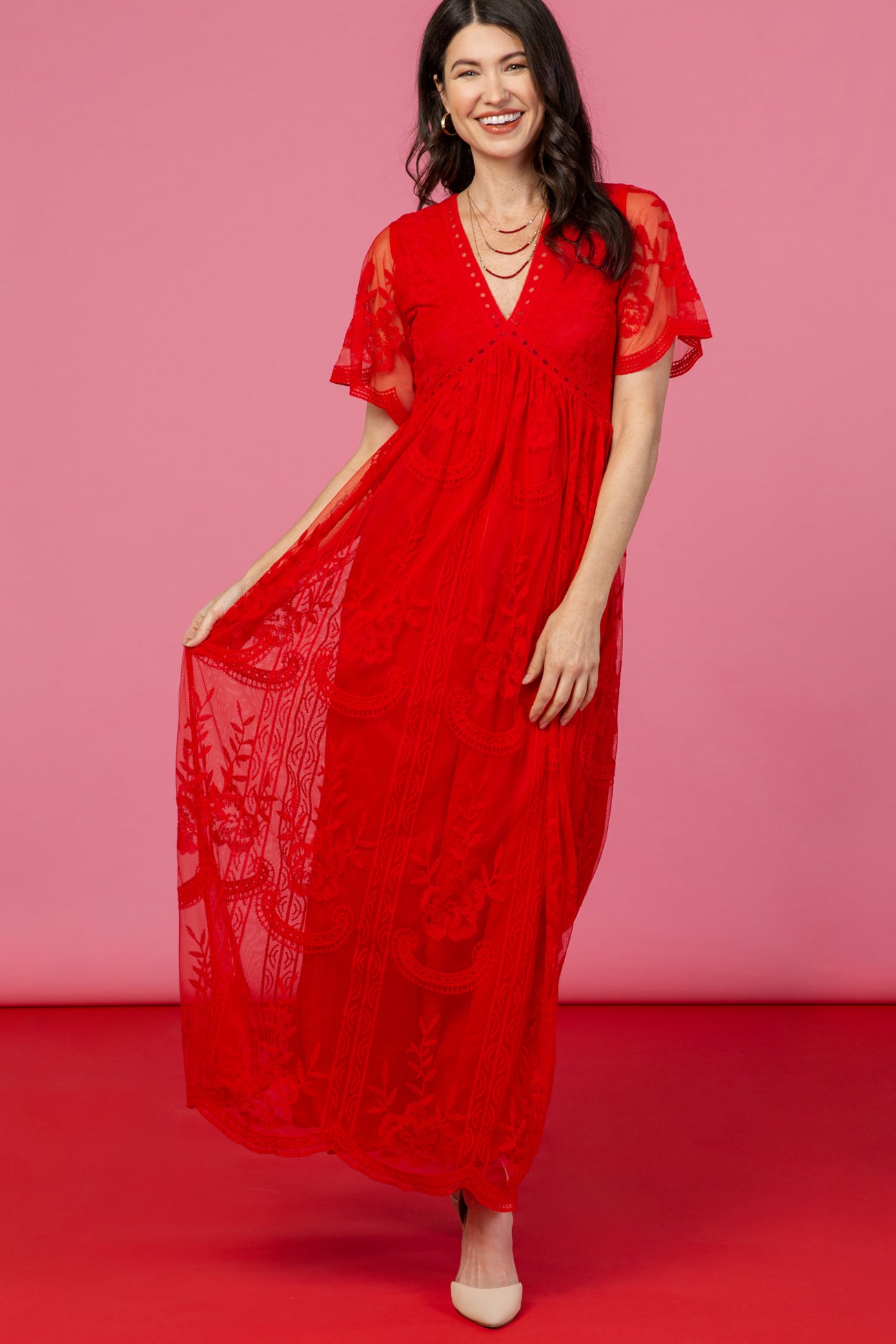 woman wearing a red lace maxi dress with short sleeves and floral pattern, smiling against a pink background
