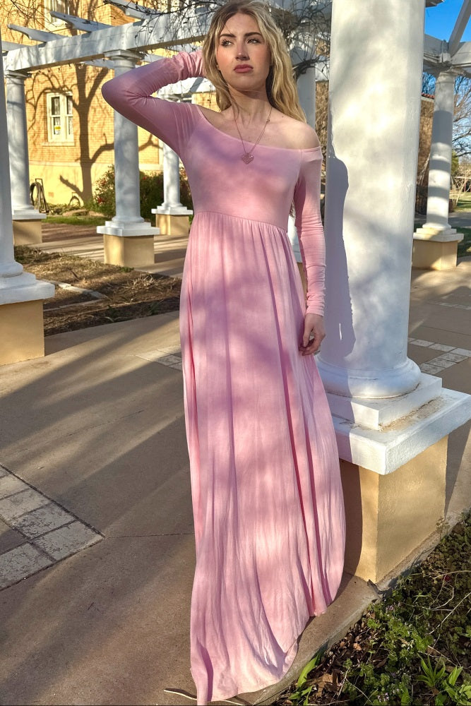 woman wearing a soft pink off-shoulder long sleeve maxi dress, standing outdoors near columns and greenery