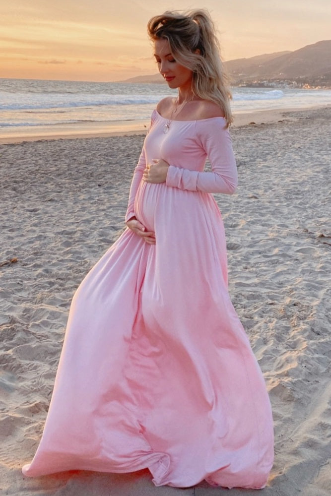 woman wearing a long pink maternity dress, standing on the beach and cradling her baby bump