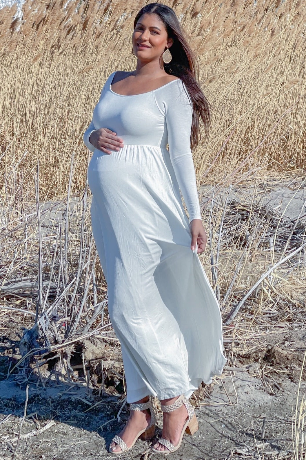 woman wearing a long white maternity dress with a fitted bodice and flowing skirt, holding her baby bump outdoors