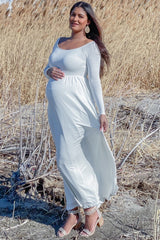 woman wearing a long white maternity dress with a fitted bodice and flowing skirt, holding her baby bump outdoors