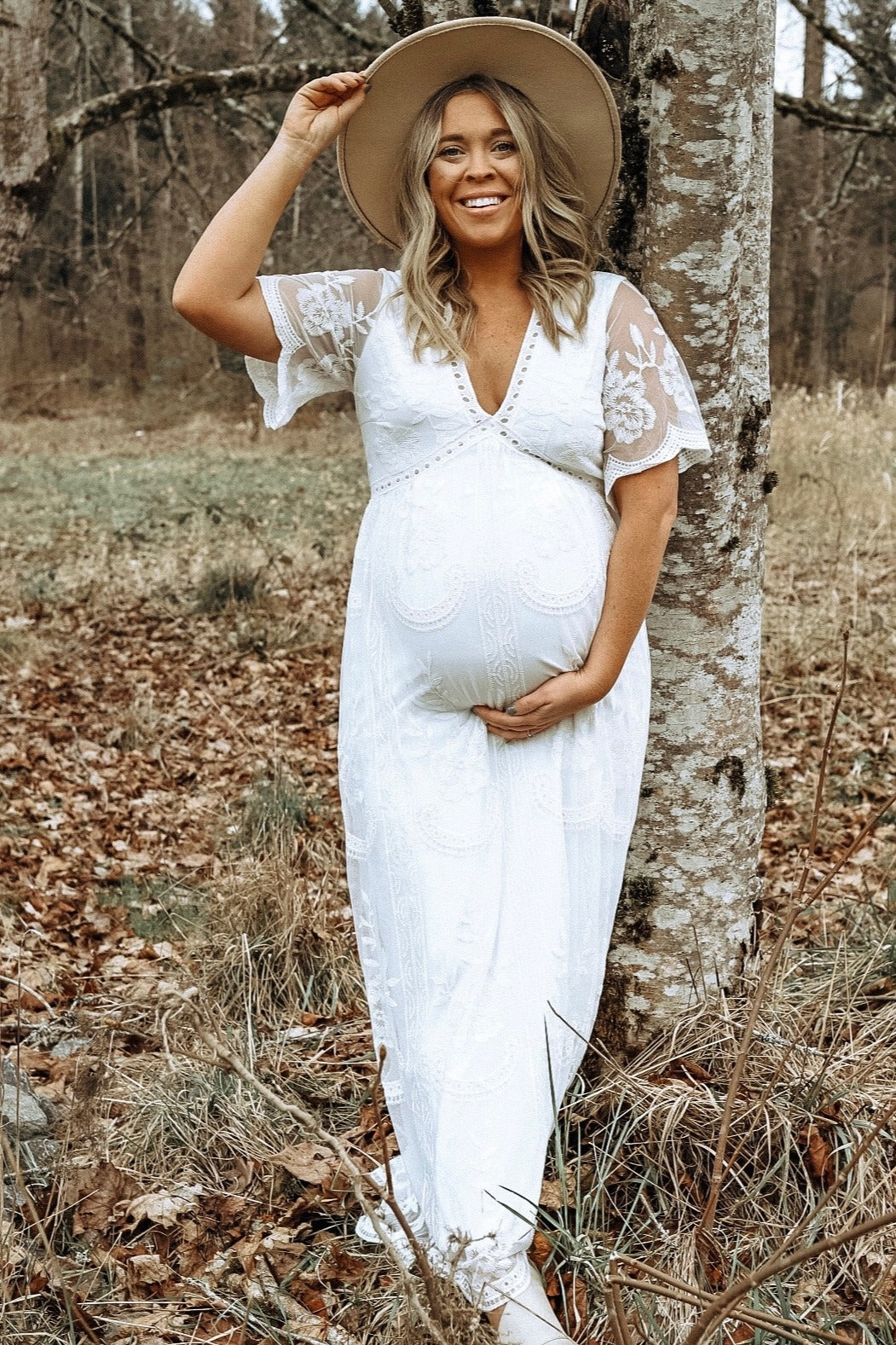 woman wearing a white lace maternity dress and holding her baby bump while smiling outdoors.