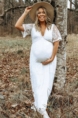 woman wearing a white lace maternity dress and holding her baby bump while smiling outdoors.