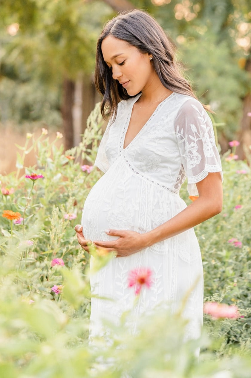 woman wearing a white lace maternity dress holding her baby bump in a floral garden