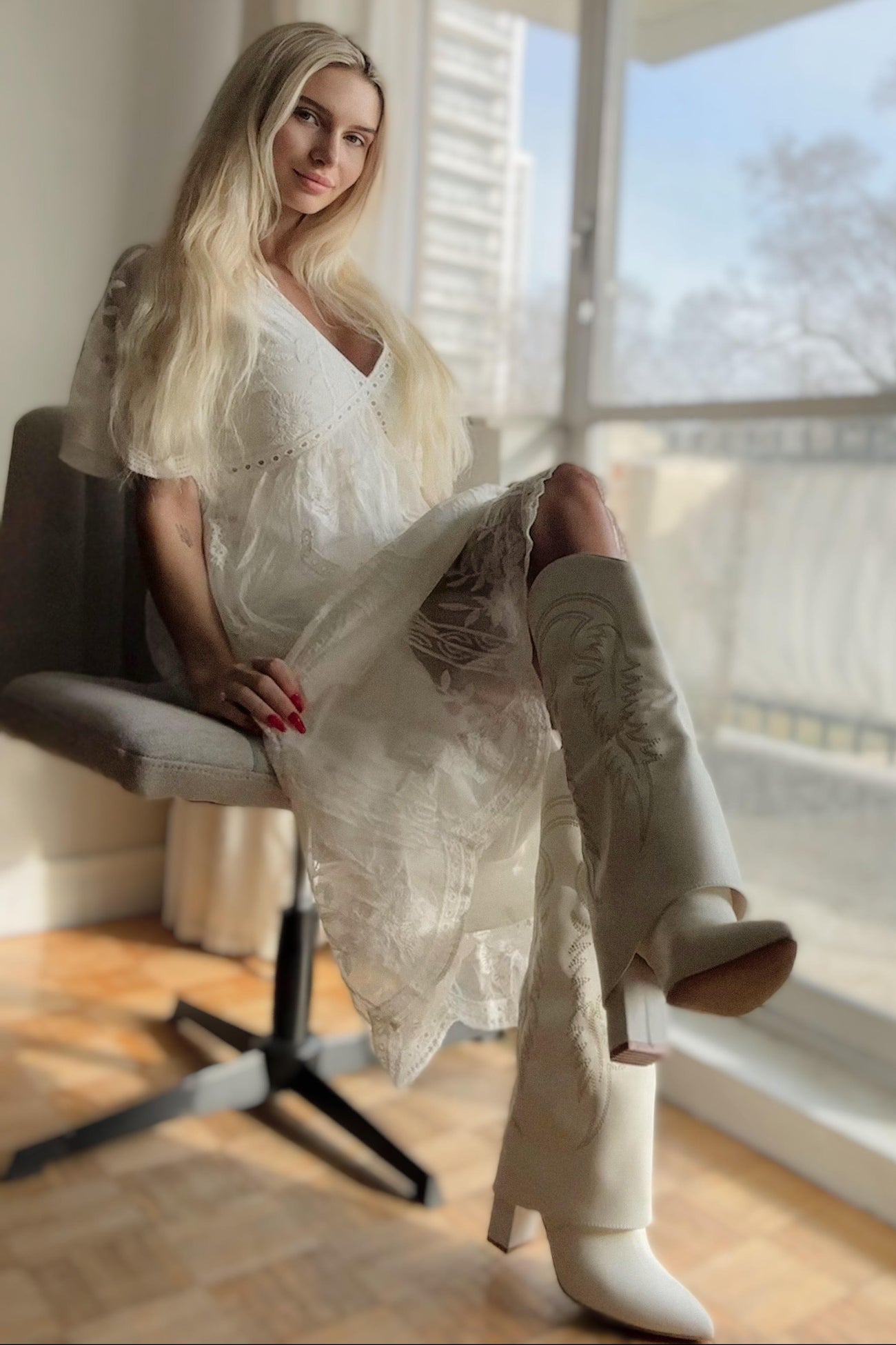 woman wearing a white lace maternity dress sitting in a chair, showcasing knee-high white heeled boots and a visible baby bump