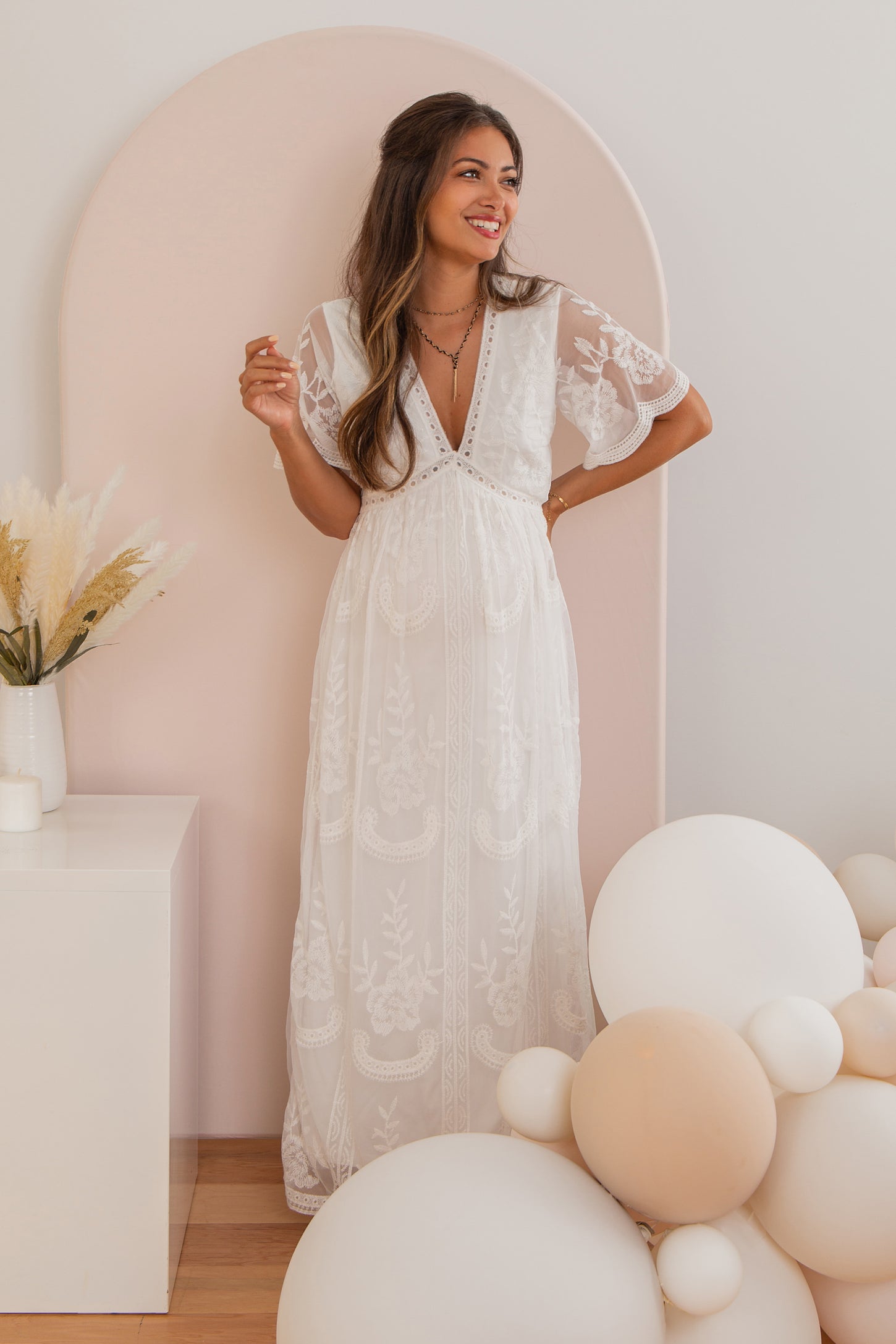 woman wearing a white embroidered maternity dress with short flutter sleeves and a v-neck, showing her baby bump