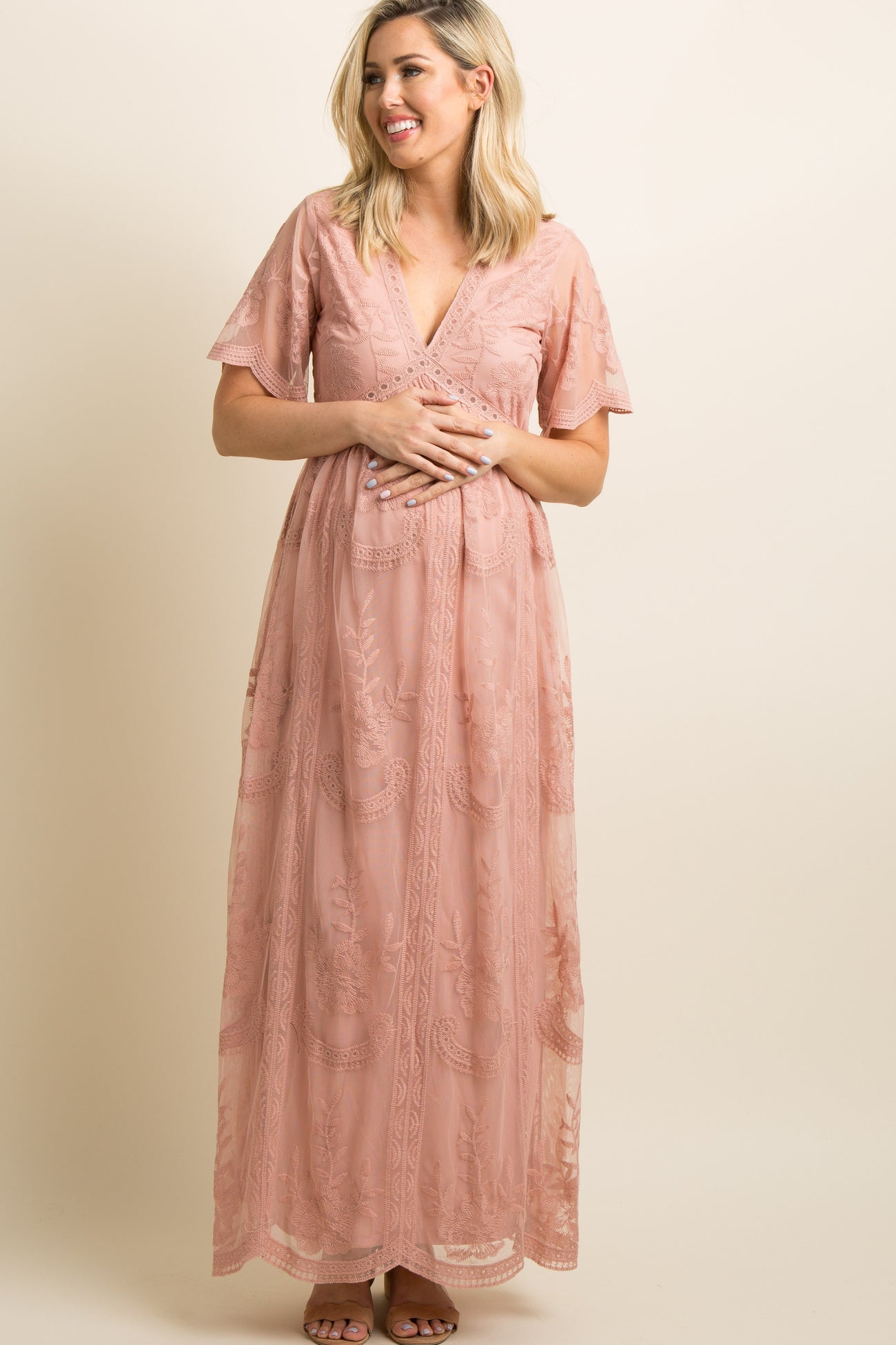 woman wearing a light peach maternity gown with floral embroidery and short flutter sleeves, showcasing her baby bump
