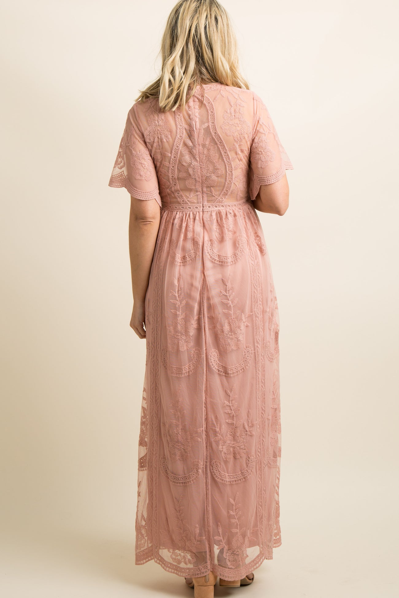 woman wearing a flowing light pink maternity gown with floral embroidery and holding her baby bump in a grassy field