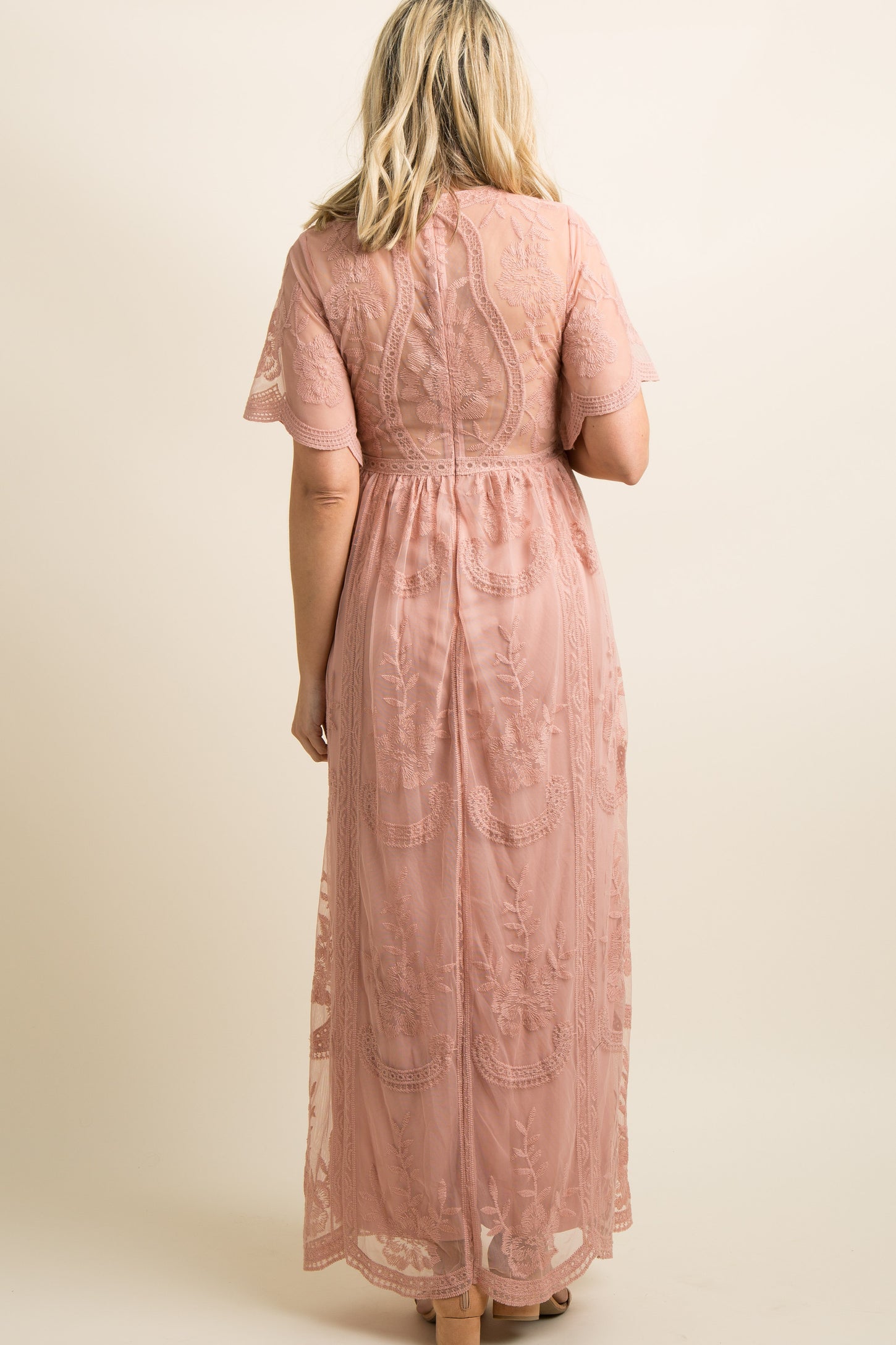 woman wearing a flowing light pink maternity gown with floral embroidery and holding her baby bump in a grassy field