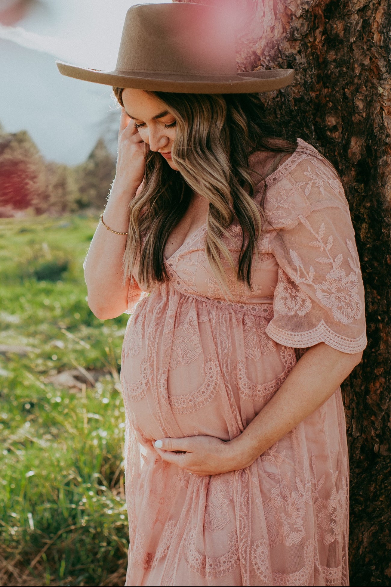 woman wearing a blush pink lace maternity dress, cradling her baby bump under a wide-brimmed hat