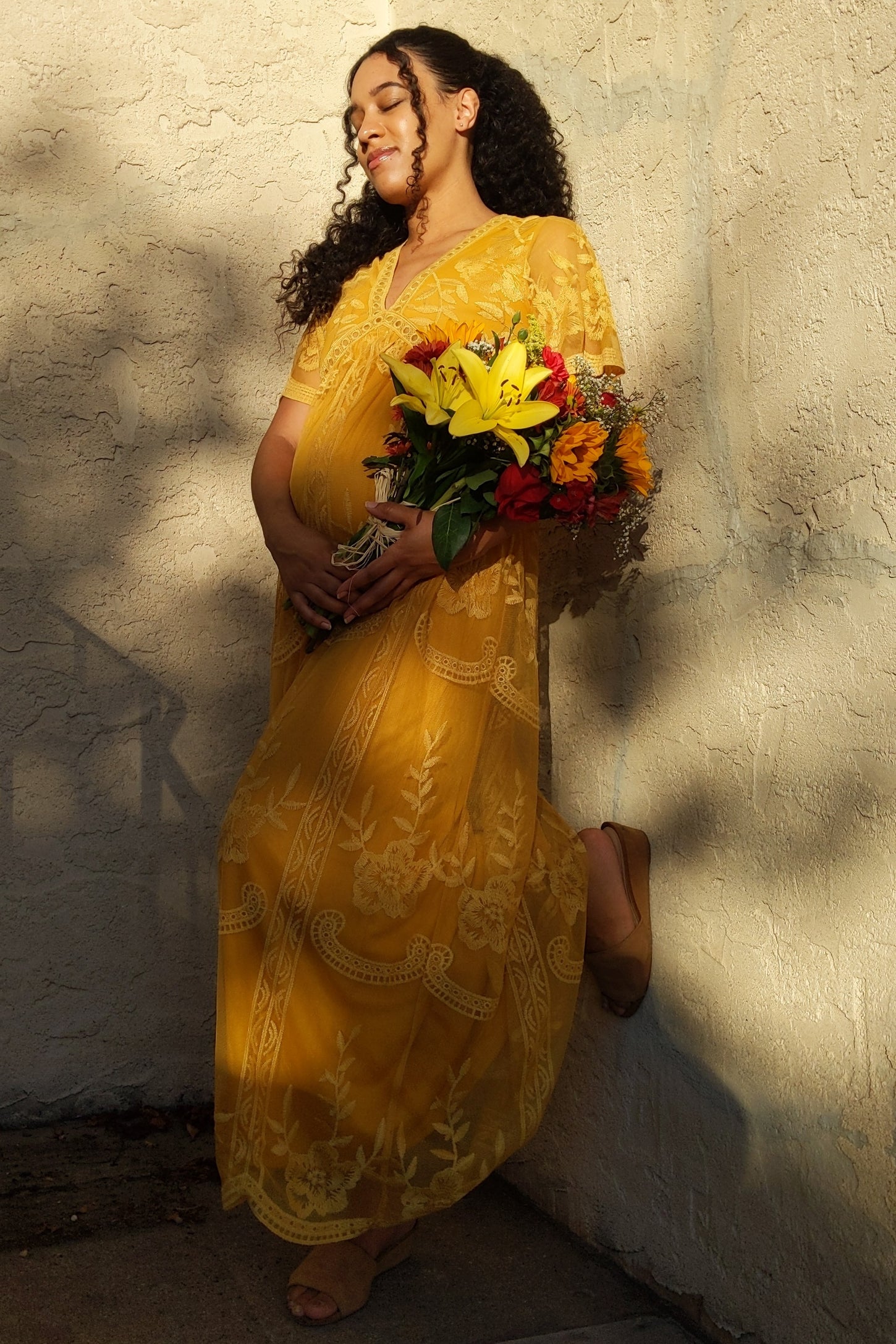 woman wearing a yellow maternity dress with floral embroidery, holding a bouquet, and showing her baby bump