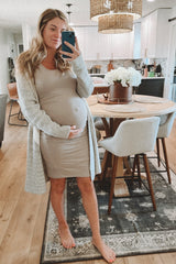 woman wearing a beige maternity dress and a light gray cardigan, cradling her baby bump in a cozy home setting