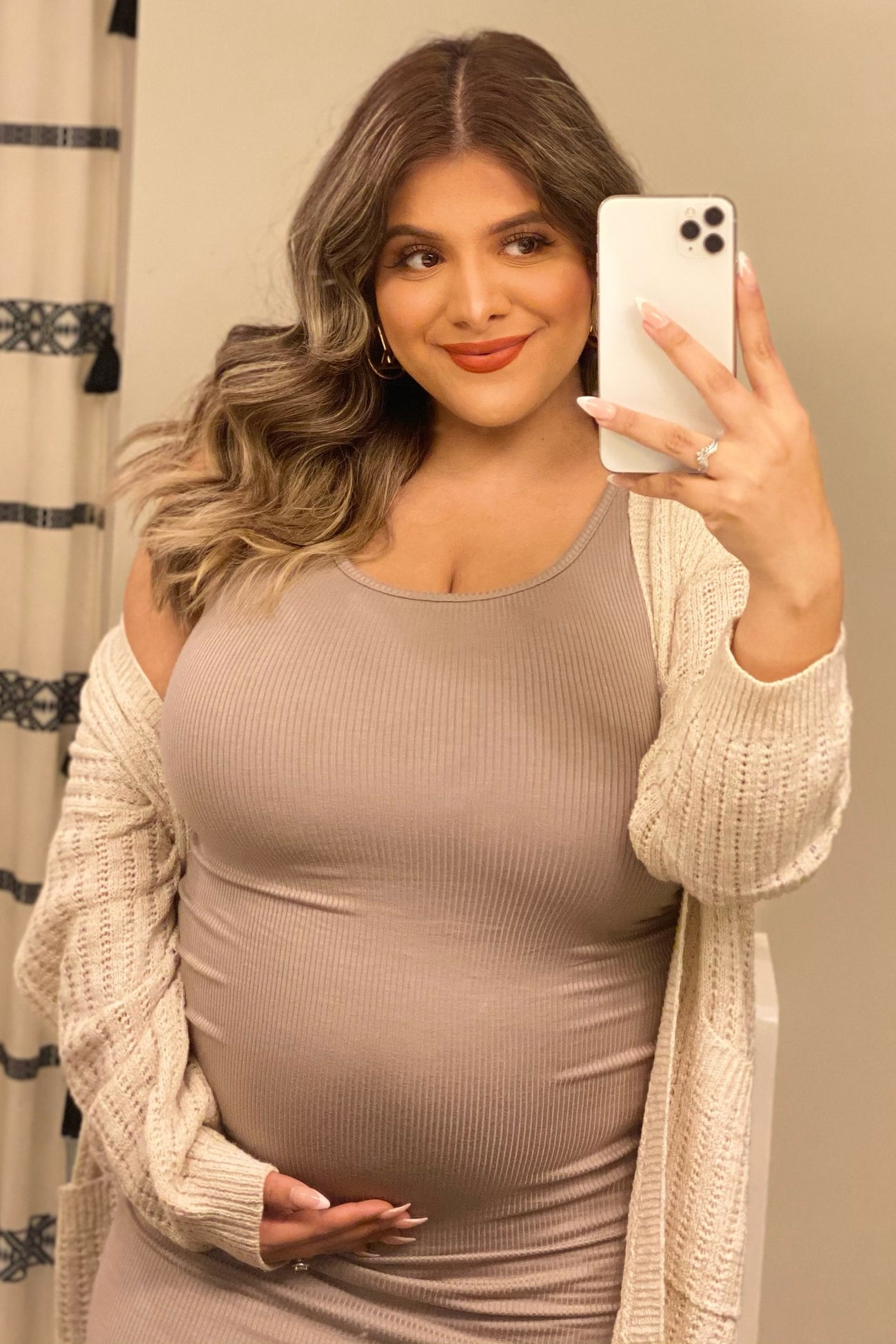 woman wearing a beige ribbed maternity dress and holding her baby bump, with a cream cardigan and styled hair