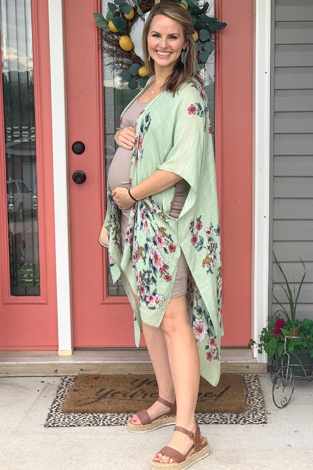 woman wearing a light green floral maternity kimono over a beige dress, showcasing her baby bump while standing by a door