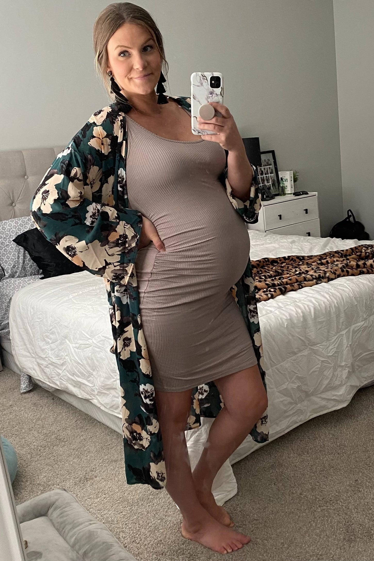 woman wearing a beige maternity dress with a floral kimono, showing her baby bump while posing in a bedroom
